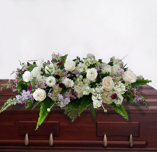 Casket spray featuring white roses, ranunculus, lilac, clematis, stock, hydrangeas, fern and more.