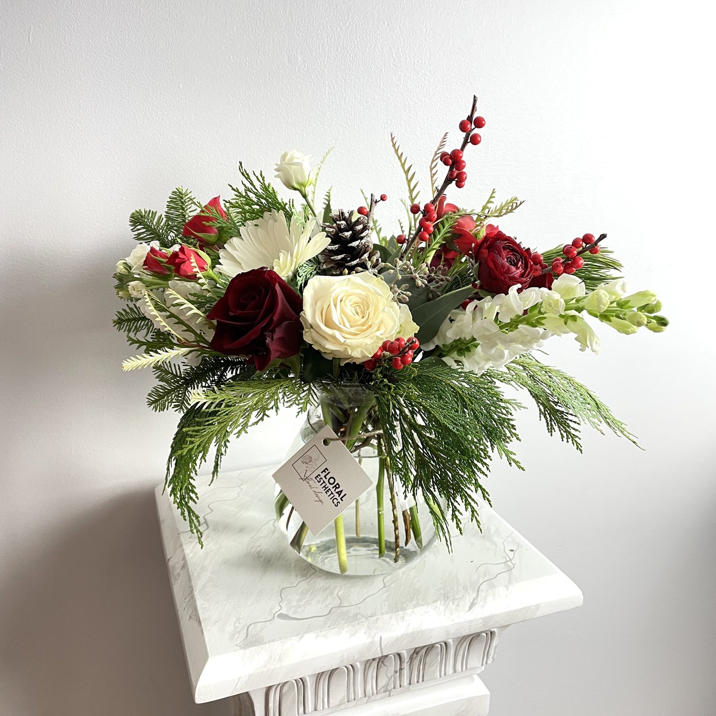 Merry Christmas Arrangement #3 featuring seasonal greens and blooms such as noble fir, Incense cedar, pine tree, Ilex berries, pine cones, roses, ranunculus, chrysanthemums and more. By Floral Esthetics