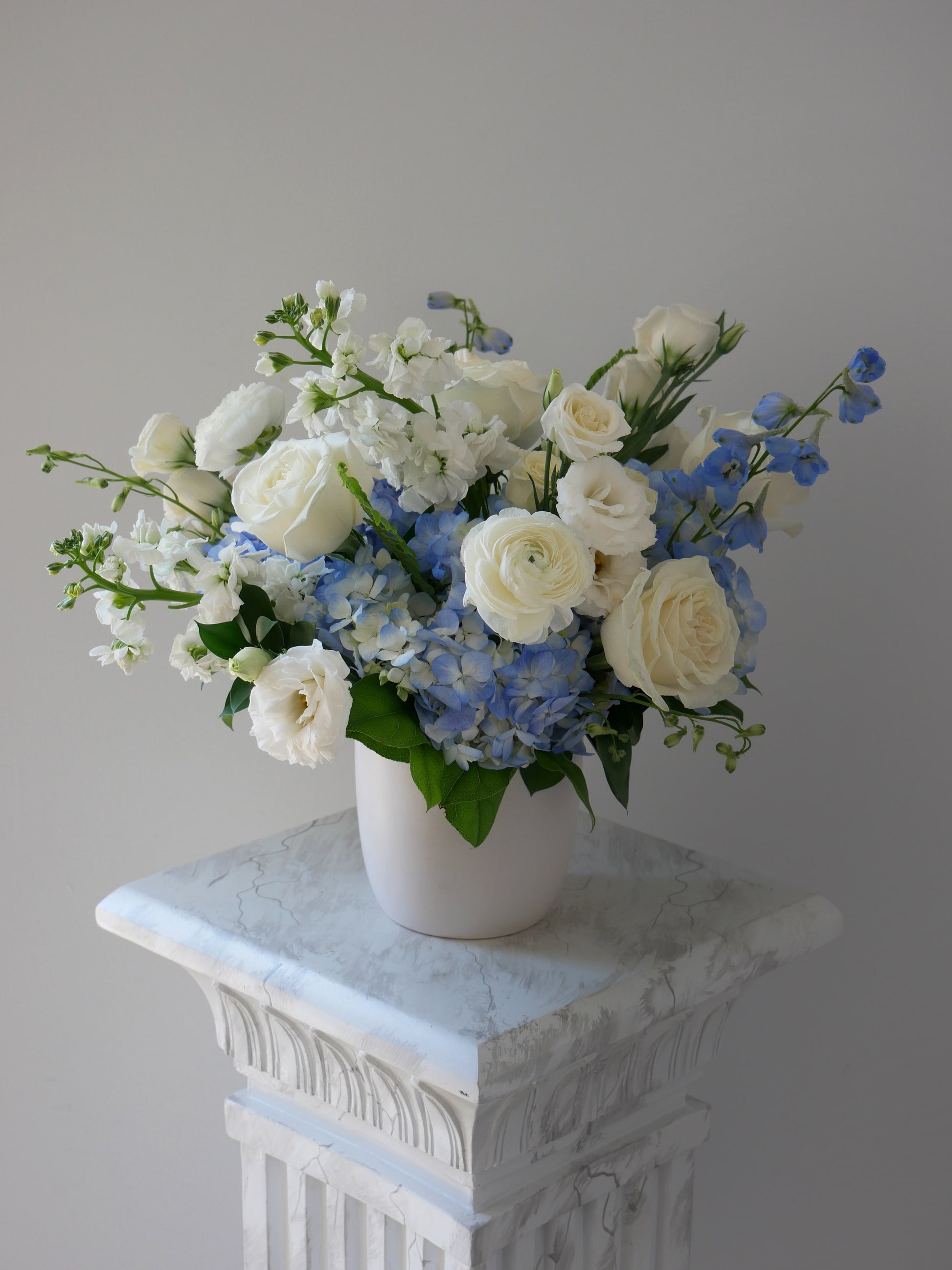 Deluxe size white and blue flower arrangement in low white cylinder featuring hydrangea, ranunculus, roses, delphinium, lizianthus, rich greens and more