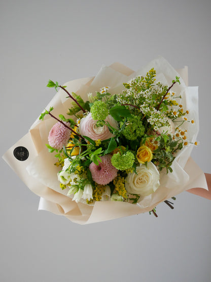 Wrapped bouquet in garden style featuring light pink ranunculus, chrysanthemum, white roses, yellow spray roses, viburnum and more