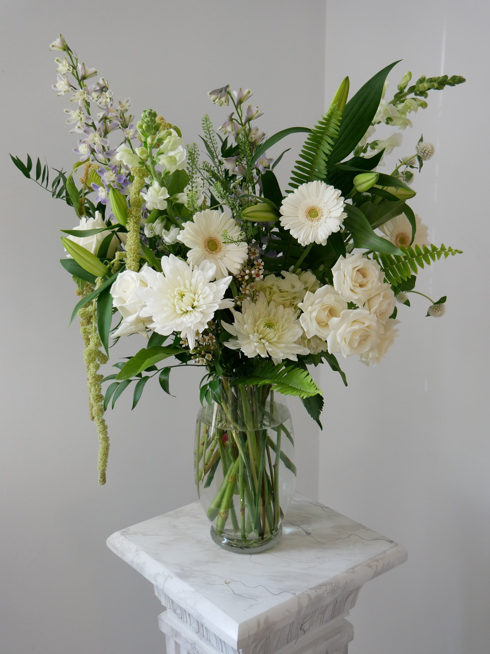 White tall flower arrangement in clear vase featuring gerberas, chrysanthemum, roses, delphinium, snap dragon, fern and much more