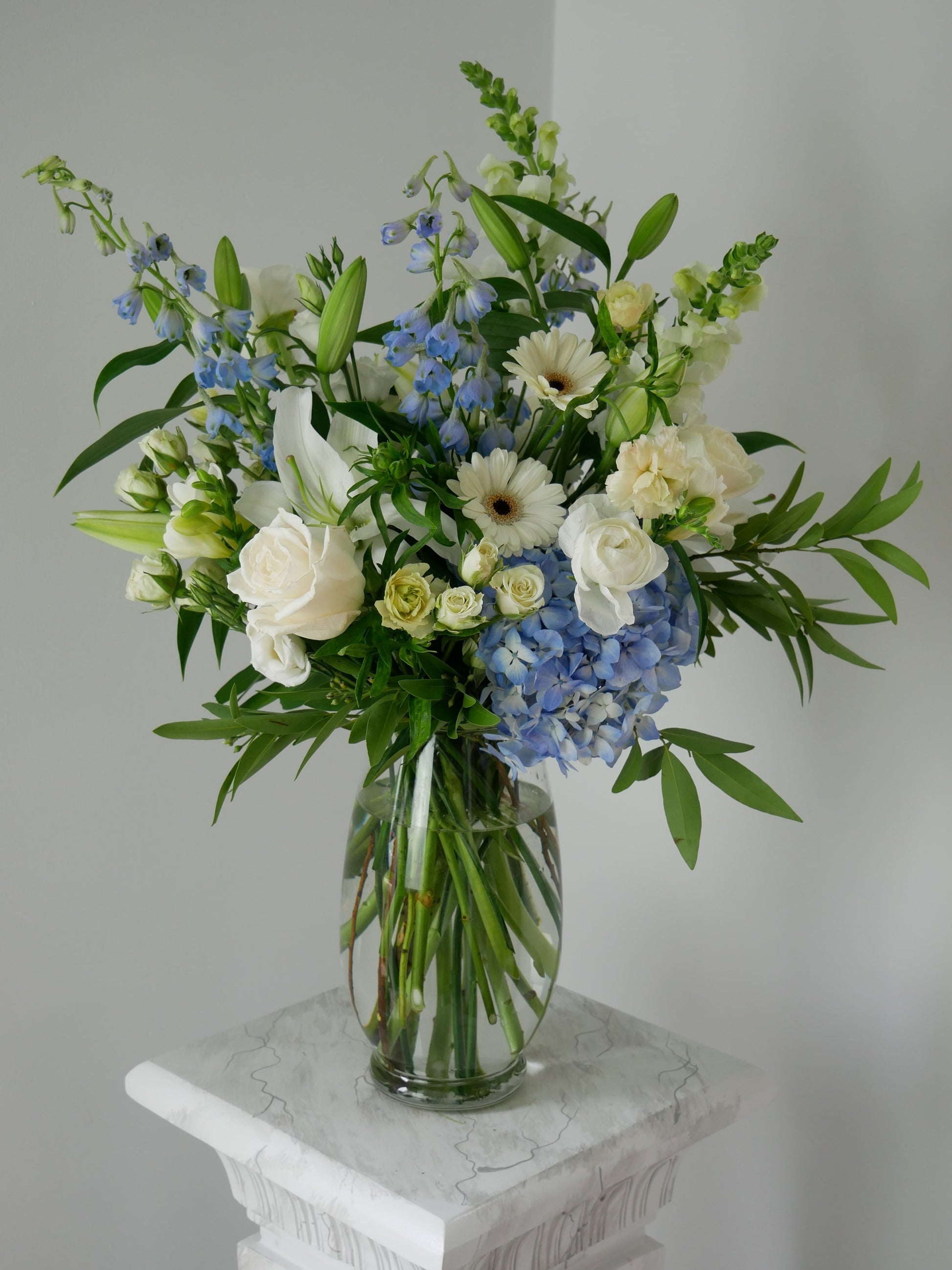 Different side of Tall pastel color flower arrangement in clear vase featuring hydrangeas, lilies, snap dragon, delphinium, ranunculus and more