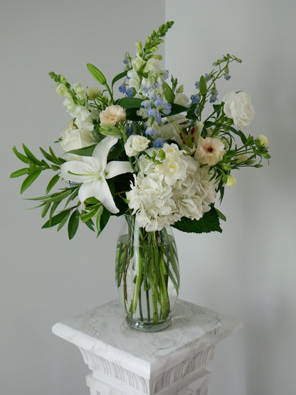 Tall pastel color flower arrangement in clear vase featuring hydrangeas, lilies, snap dragon, delphinium, ranunculus and more