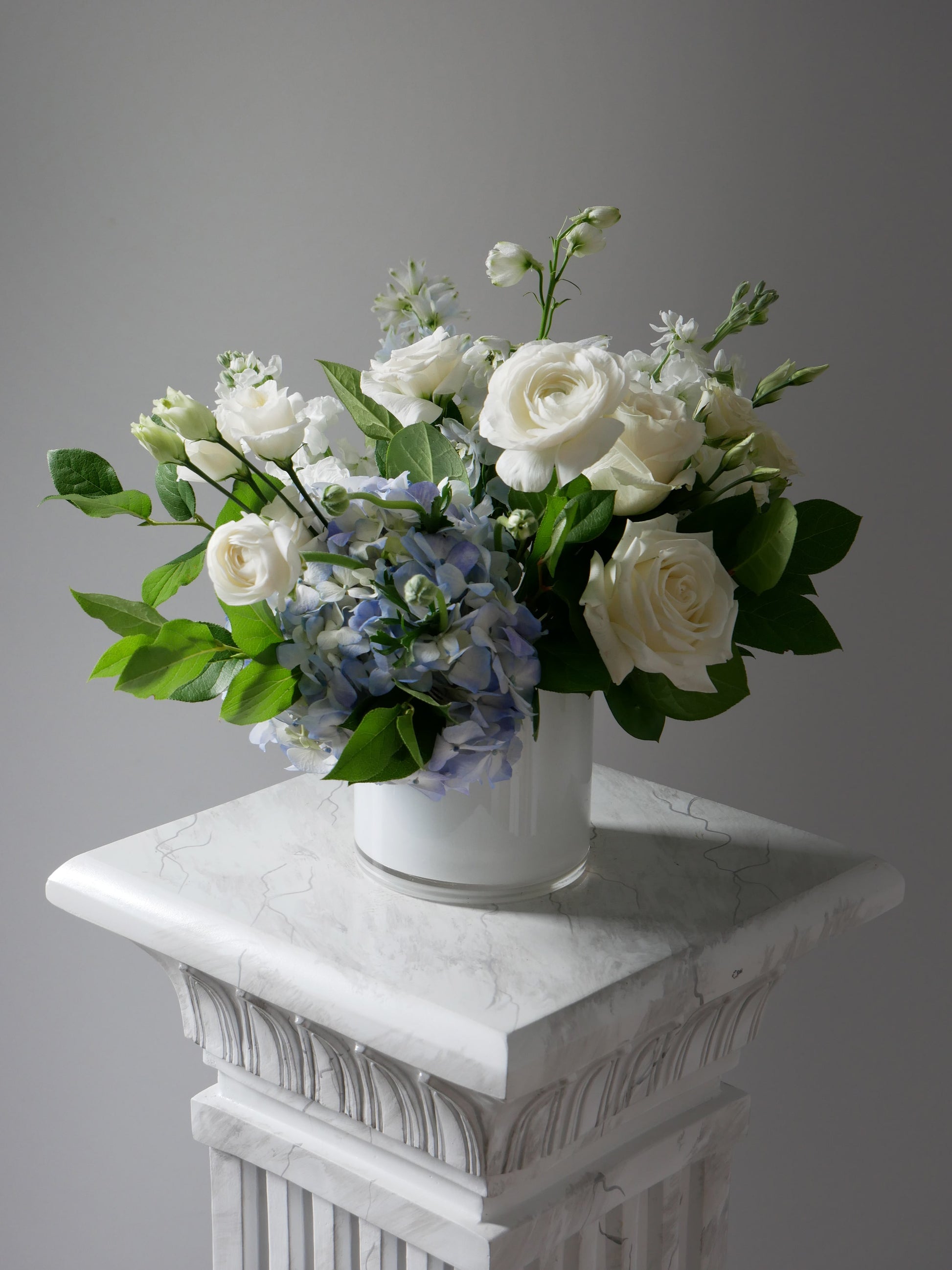 White and blue flower arrangement in low white cylinder featuring hydrangea, ranunculus, roses, delphinium, lizianthus, rich greens and more