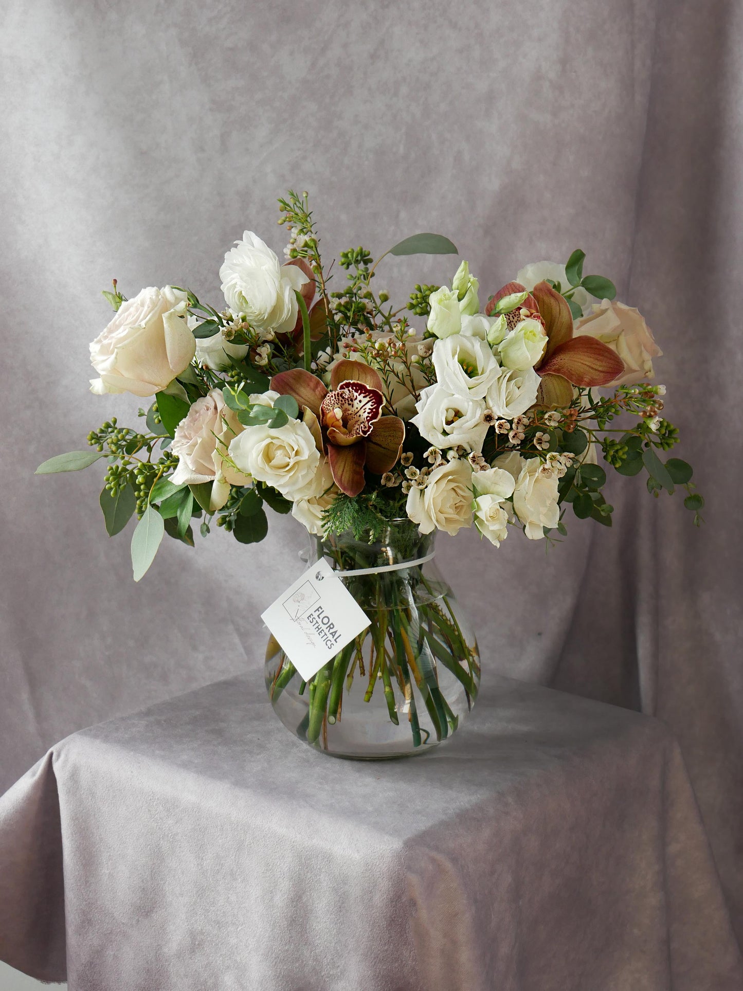 White and coffee color arrangement in clear vase featuring orchids, roses, spray roses, lizianthus, eucalyptus, wax flower and more