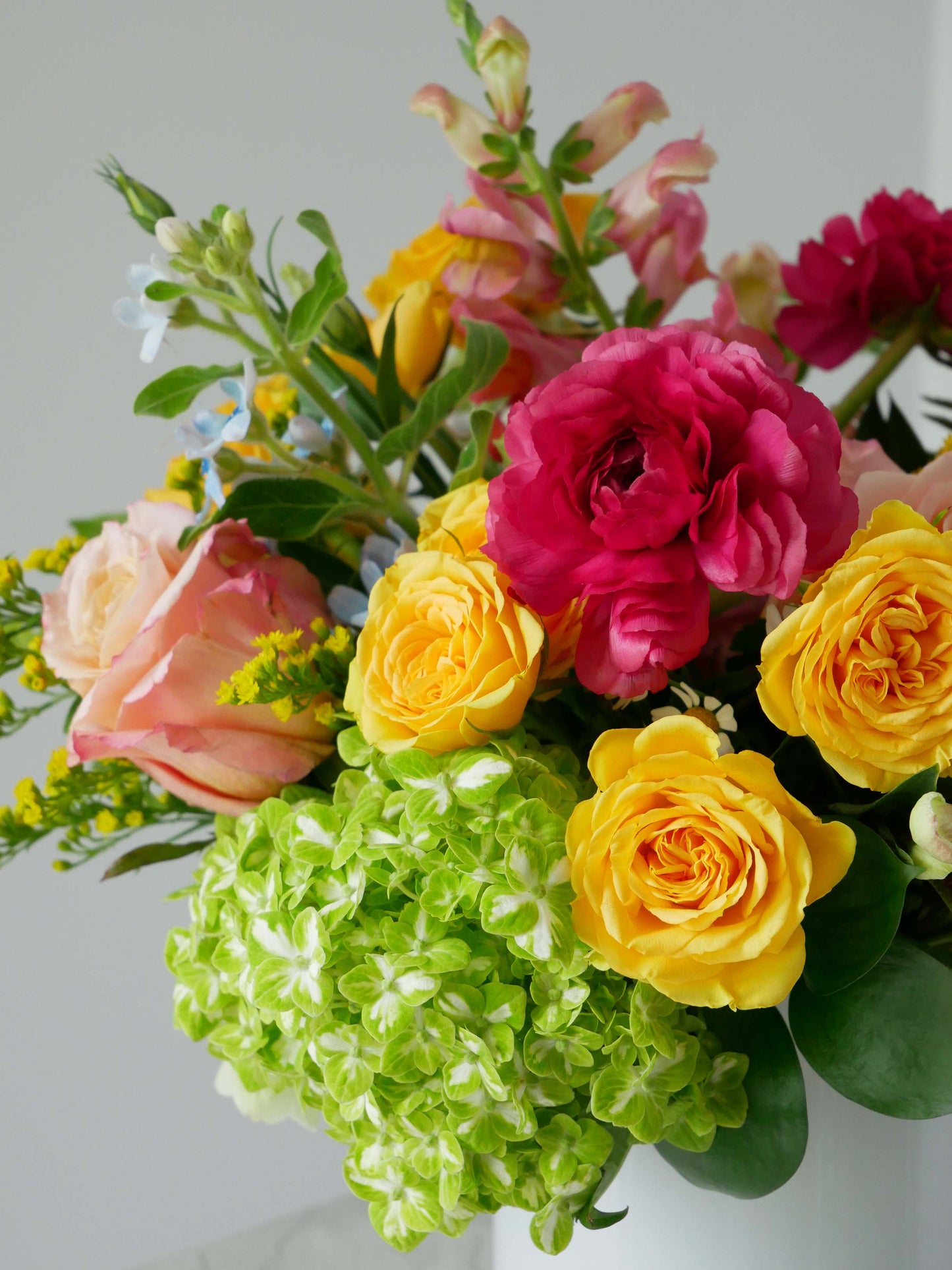 Close-up on Standard size flower arrangement in low white cylinder featuring hot pink ranunculus, yellow roses, peachy roses and greens