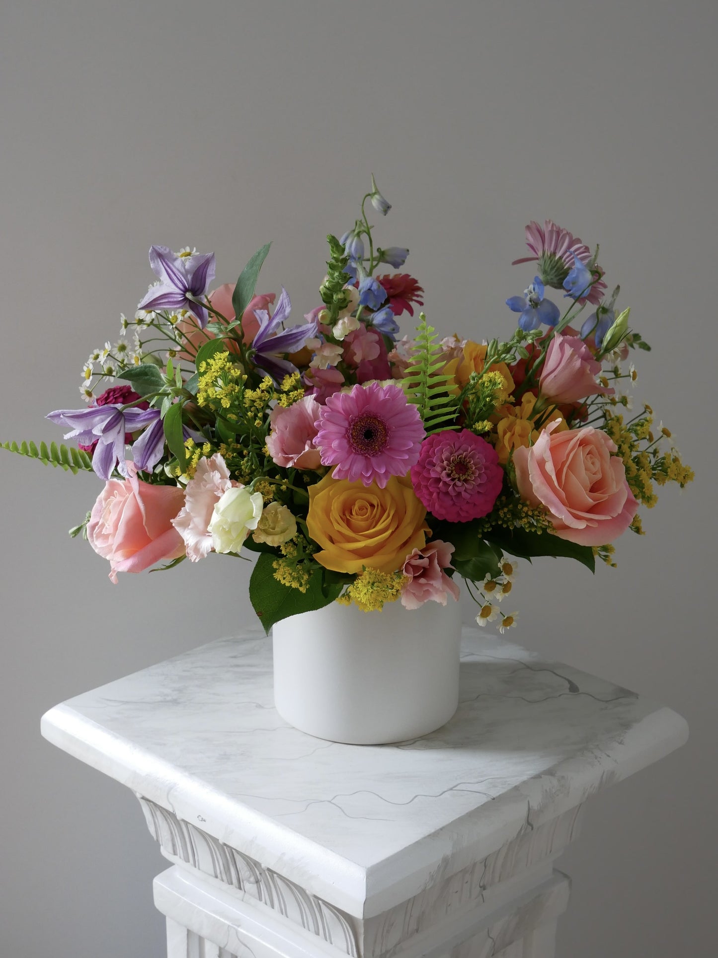 Premium size flower arrangement in low white cylinder featuring hot pink zinnia, klematis, pink lisianthus, gerberas, yellow roses,  peachy roses and greens by Floral Esthetics