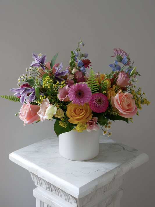 Premium size flower arrangement in low white cylinder featuring hot pink zinnia, klematis, pink lisianthus, gerberas, yellow roses,  peachy roses and greens by Floral Esthetics