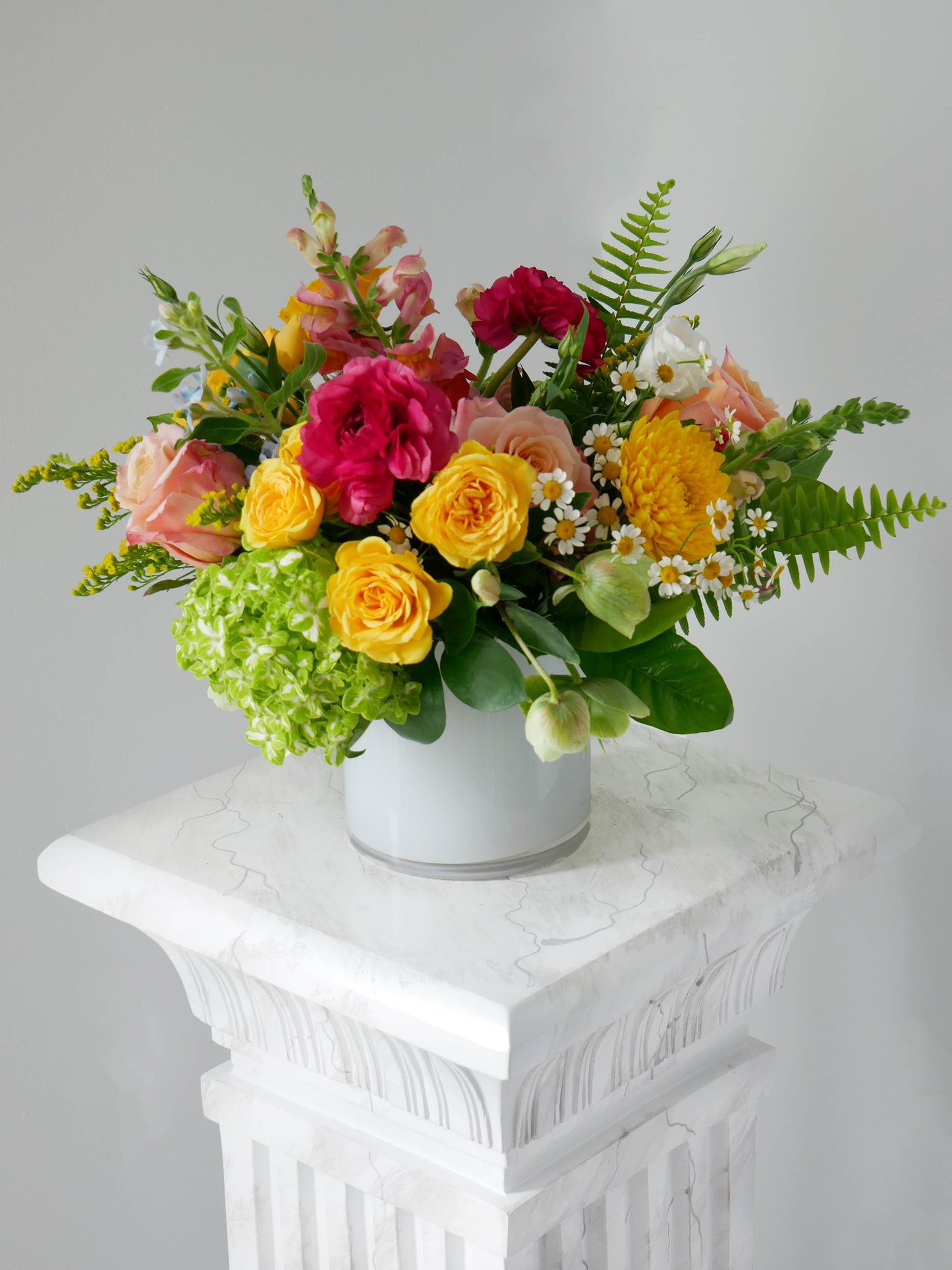 Standard size flower arrangement in low white cylinder featuring hot pink ranunculus, yellow roses, peachy roses and greens by Floral Esthetics