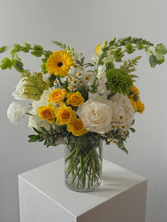 Bouquet of yellow and white flowers in a clear vase on a white surface with a gray background