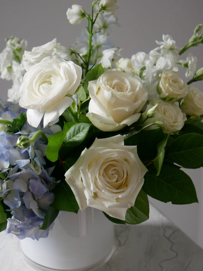 Close-up on white and blue flower arrangement in low white cylinder featuring hydrangea, ranunculus, roses, delphinium, lizianthus, rich greens and more by Floral Esthetics