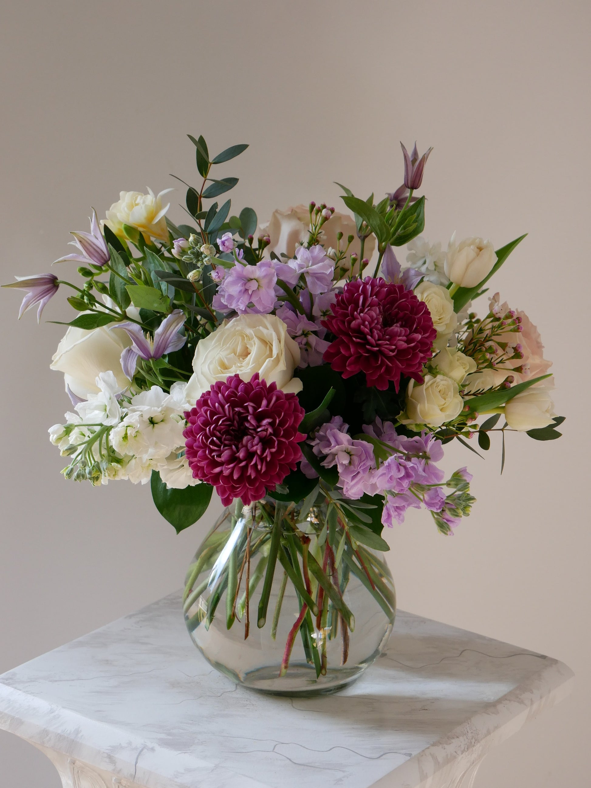 Standard size flower arrangement in glass vase n white and purple color palette featuring roses, chrysanthemum, clematis, stock and more 