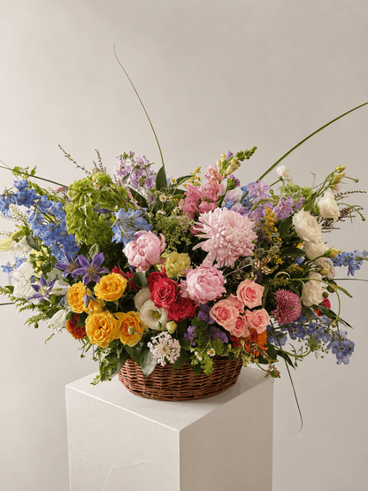 Sympathy colorful flowers in a woven basket on a white pedestal against a light gray background