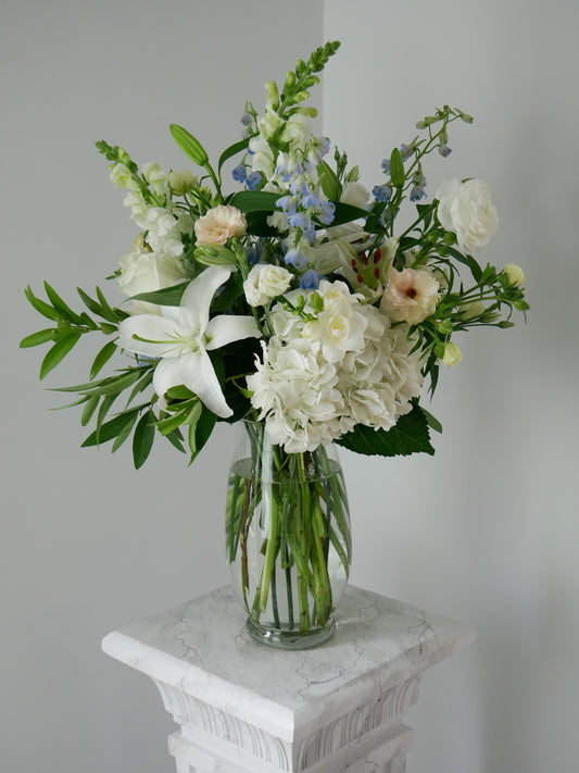 Tall pastel color flower arrangement in clear vase featuring hydrangeas, lilies, snap dragon, delphinium, ranunculus and more by Floral Esthetics