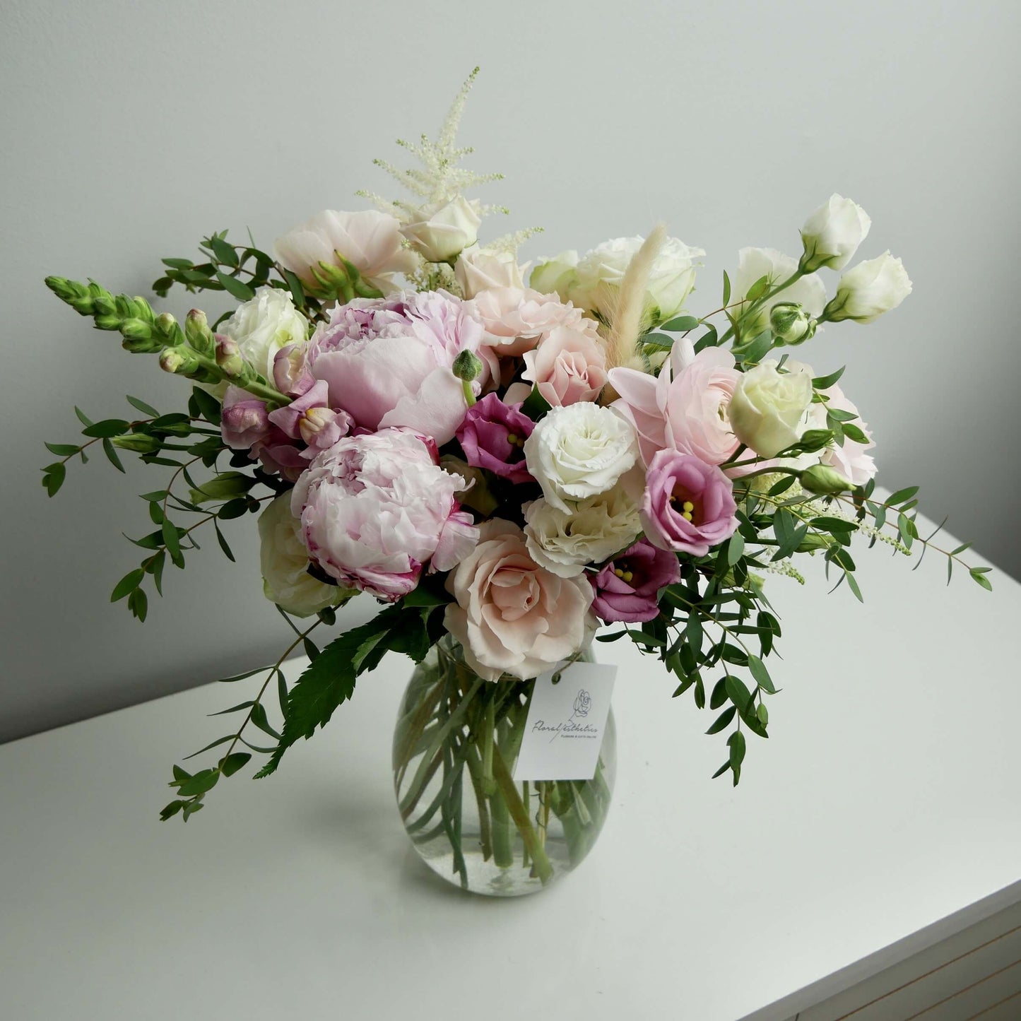 Tender pink and white standard size flower arrangement in clear vase featuring peonies, ranunculus, roses, greens and more