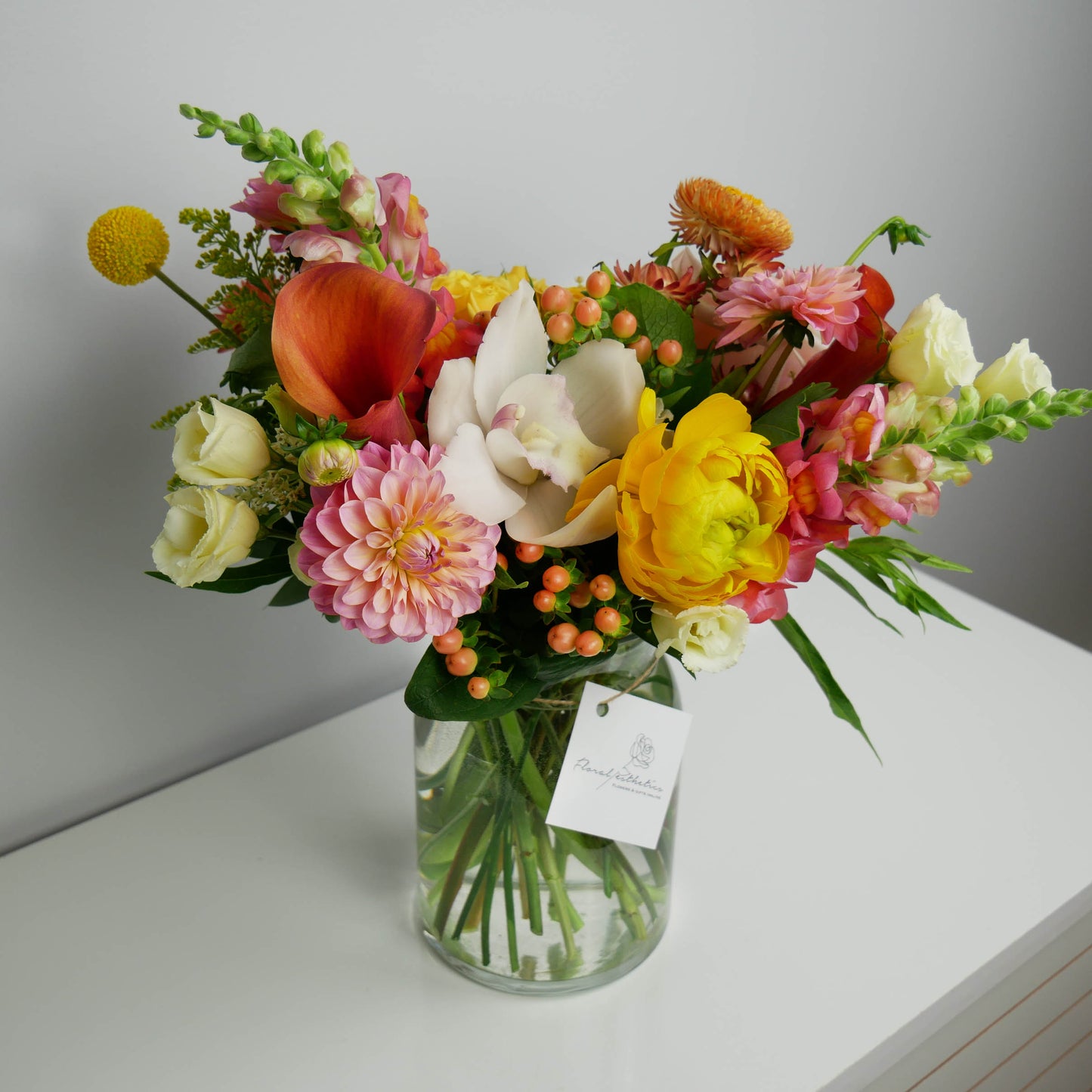 Vibrant orange pink standard size vase arrangement featuring orchids, ranunculus, snap dragon, dahlia and more
