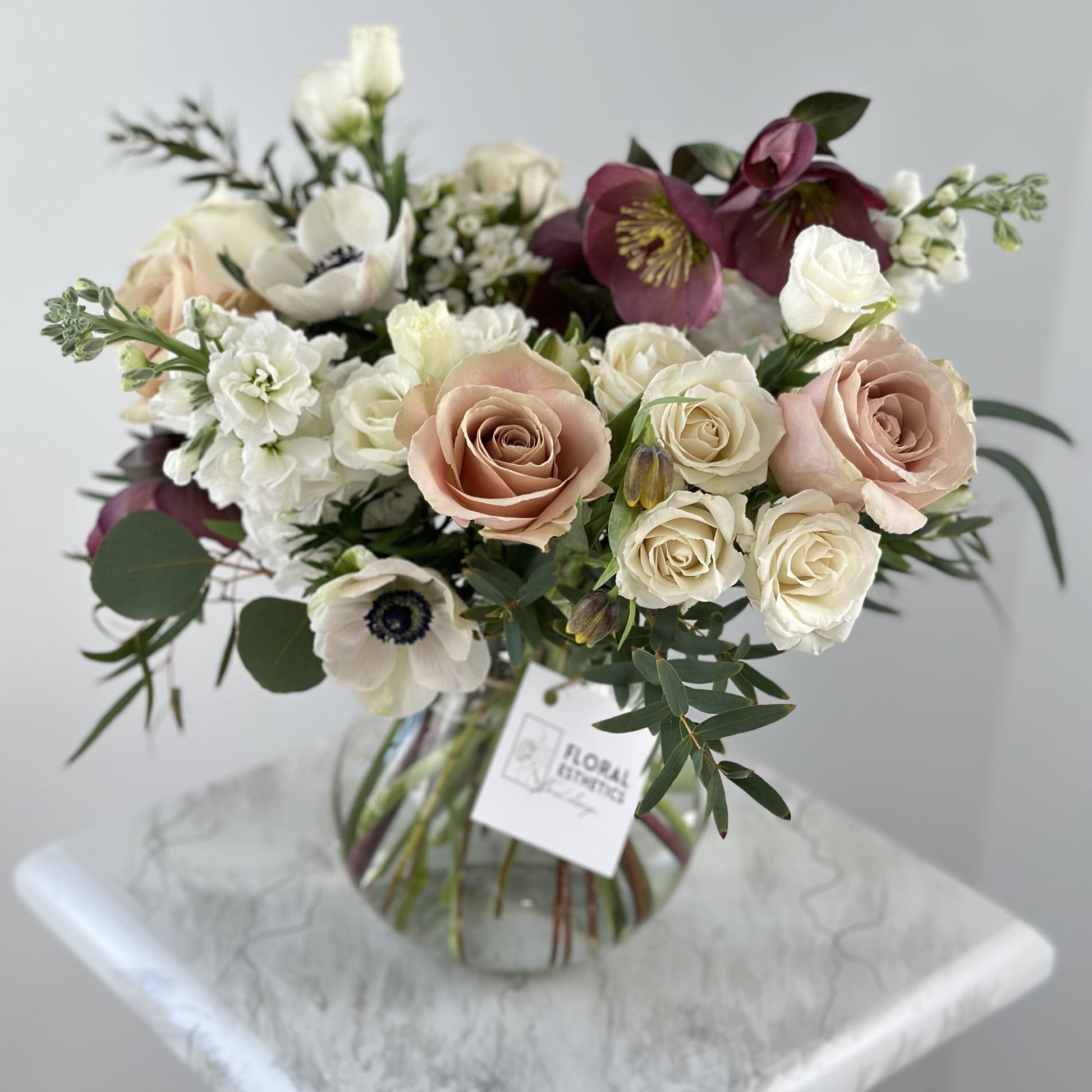 White and coffee color arrangement in clear vase featuring anemone, roses, spray roses, lizianthus, hellebore, eucalyptus, wax flower and more