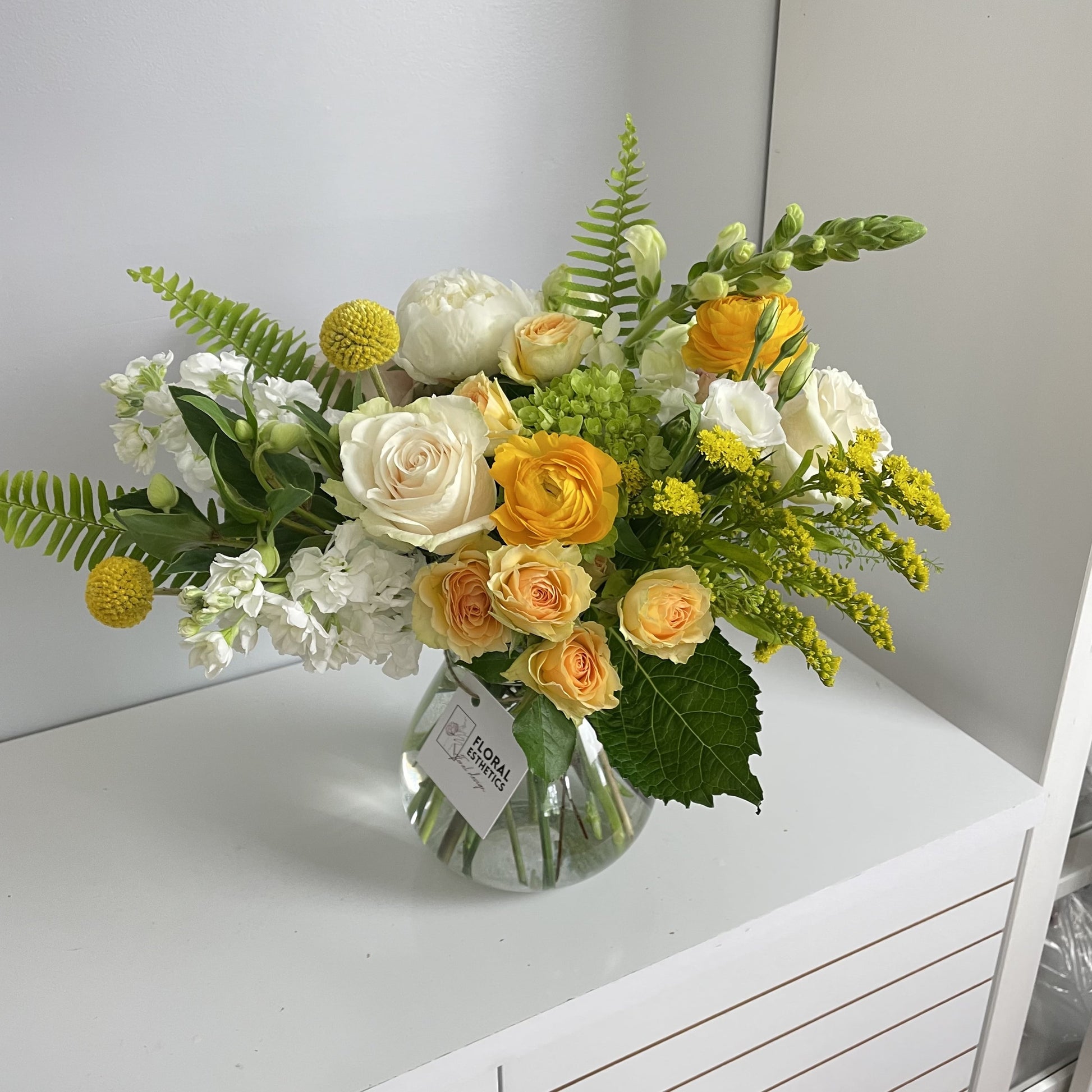 White and yellow flower arrangement in clear vase featuring ranunculus, roses, hydrangeas, greens and more