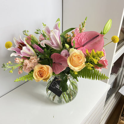 Colorful tropical flower arrangement in clear vase featuring lilies, anthurium, roses, snap, craspedia and much more