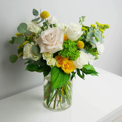 White and yellow flower arrangement in clear vase featuring roses, hydrangeas, greens and more