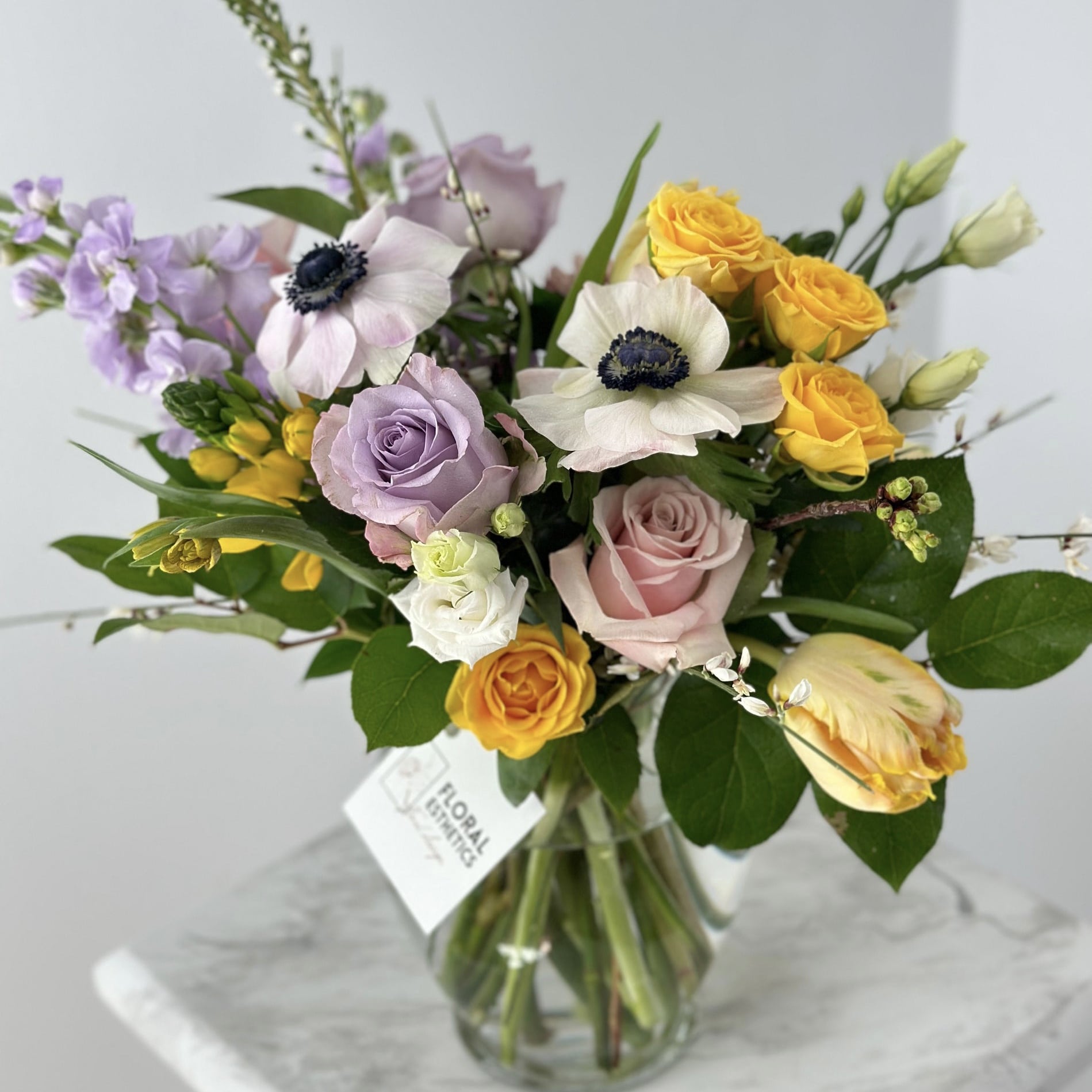 Spring flower arrangement in clear vase featuring white anemones, purple roses, yellow spray roses, yellow parrot tulips, genistra, freesia and more