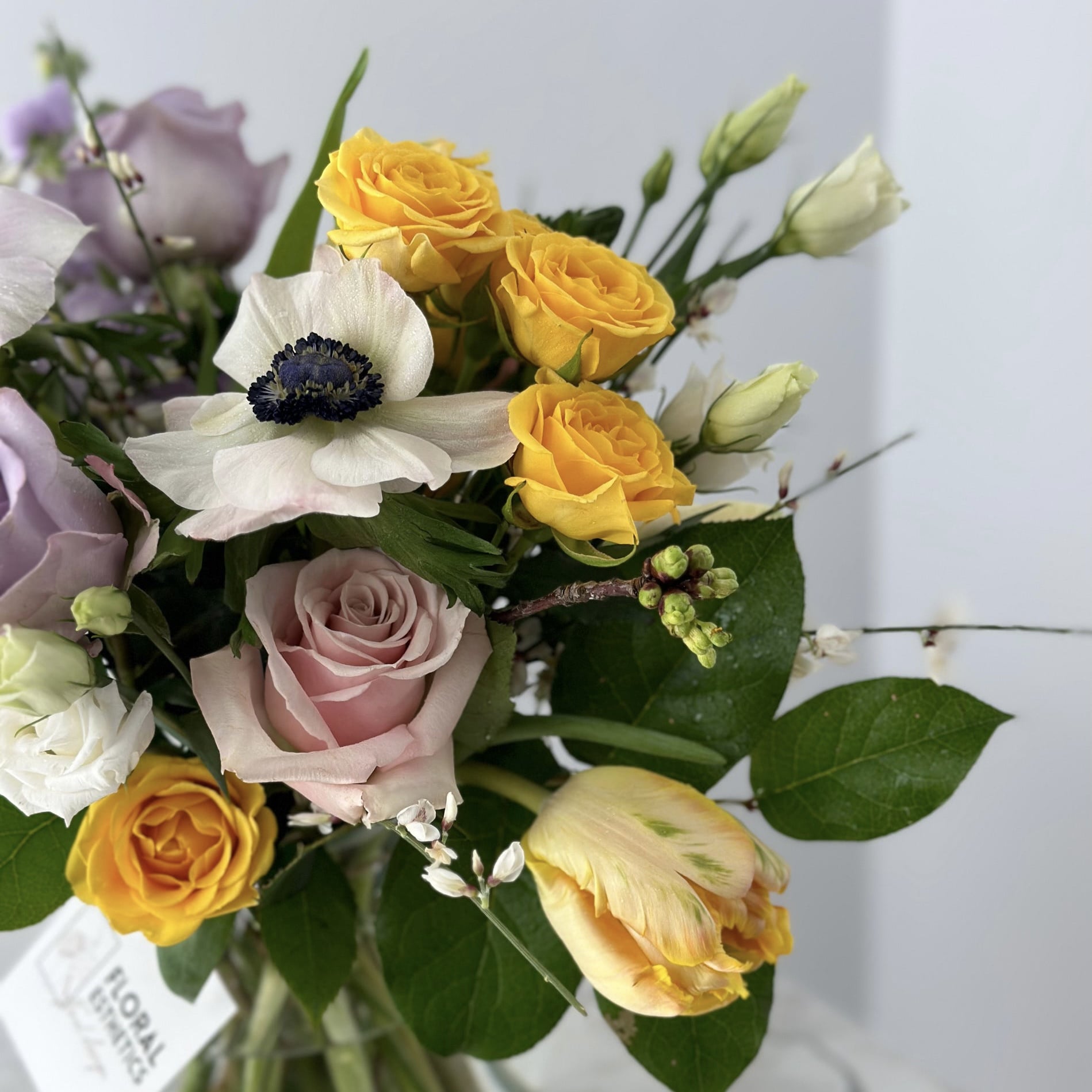 Close-up on Spring flower arrangement in clear vase featuring white anemones, purple roses, yellow spray roses, yellow parrot tulips, genistra, freesia and more