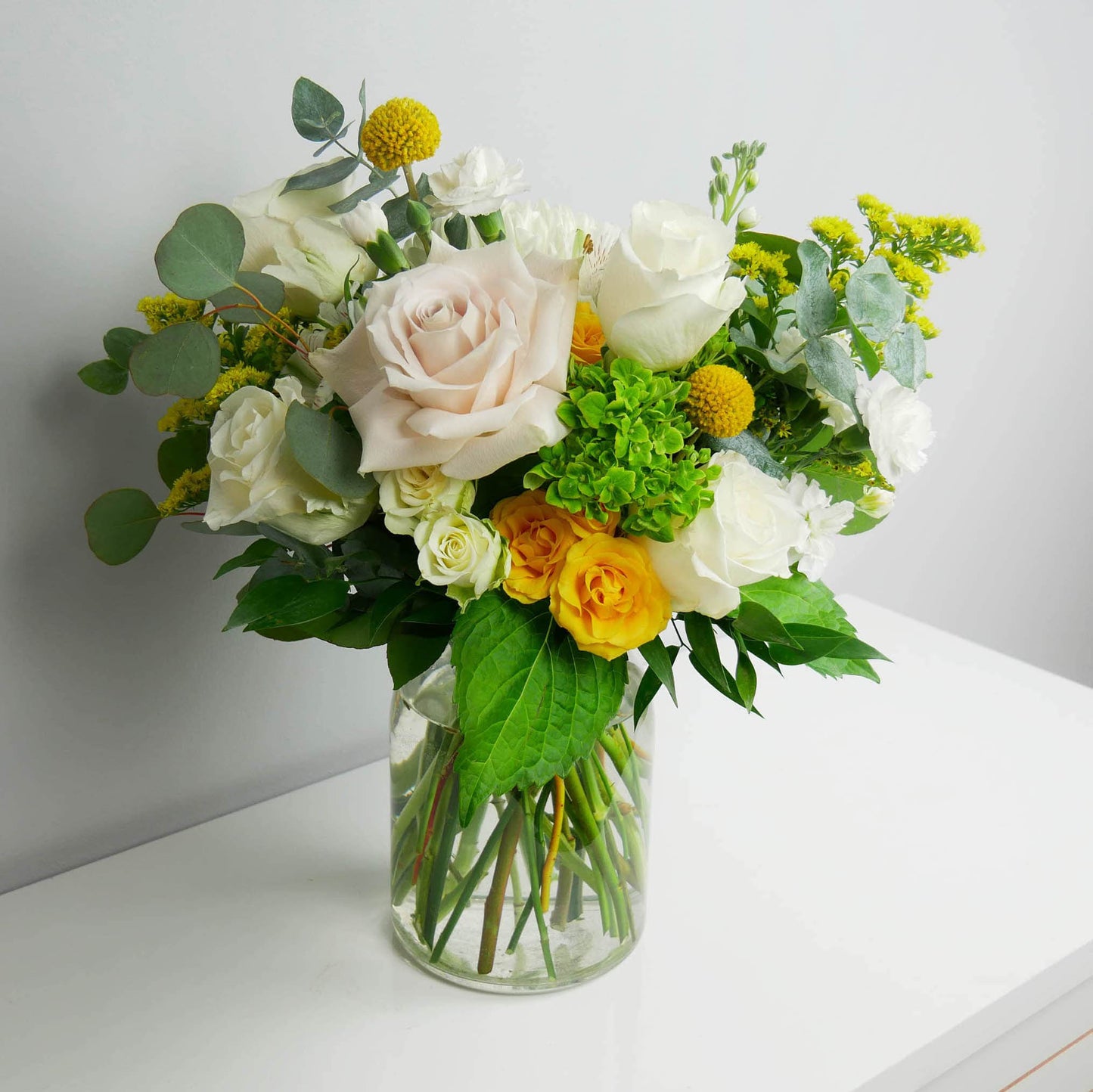 White and yellow flower arrangement in clear vase featuring roses, hydrangeas, greens and more by Floral Esthetics