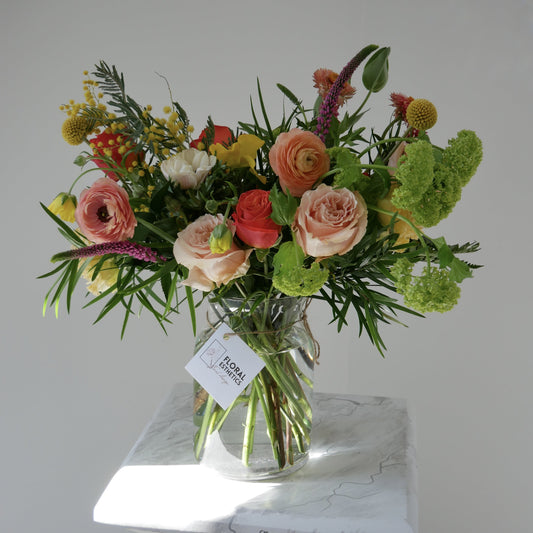 Premium size orange, yellow & green flower arrangement in clear vase featuring ranunculus, roses, viburnum, mimosa and more by Floral Esthetics